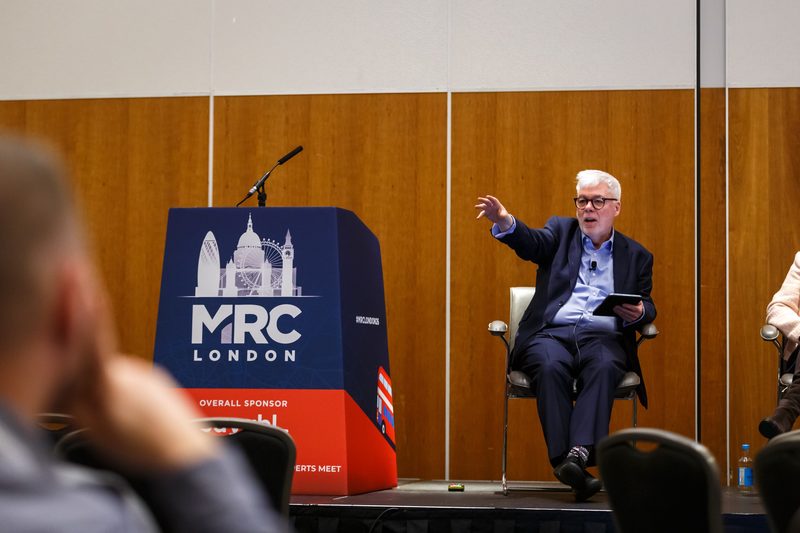 John Elliott seated on stage beside an MRC London lectern, gesturing towards the audience during a panel session.